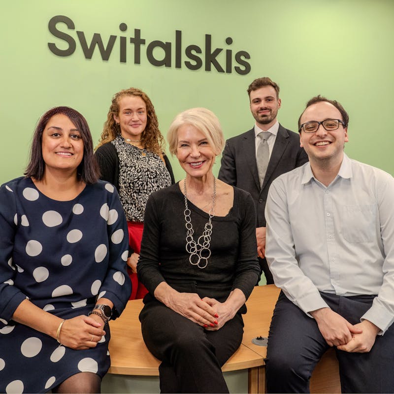 A group of five professionals is posing for a photo in an office setting. They are seated and standing in front of a light green wall with the company name "Switalskis" displayed prominently in black letters. The group consists of three women and two men. The woman on the left is wearing a navy blue dress with large white polka dots. Next to her is a woman with short blonde hair, wearing a black top and a long silver necklace. Standing behind them are a woman with curly red hair, a man in a dark suit and tie, and a man in glasses and a light blue shirt. They are all smiling and appear to be colleagues.